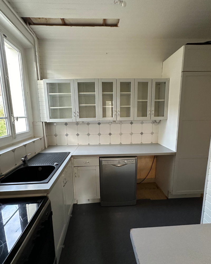HS_1_Avant_E Petite cuisine avec armoires blanches, meubles hauts vitrés, lave-vaisselle, évier noir et une porte de placard manquante, révélant un espace vide sous le comptoir. Un panneau de plafond est partiellement manquant.