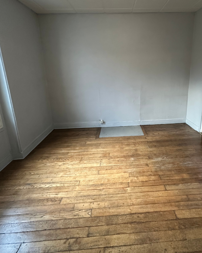 HS_1_Avant_G Une petite chambre meublée simplement avec un lit simple, une armoire en bois, un bureau, une chaise et une fenêtre avec des rideaux bleus laissant entrer la lumière du jour.