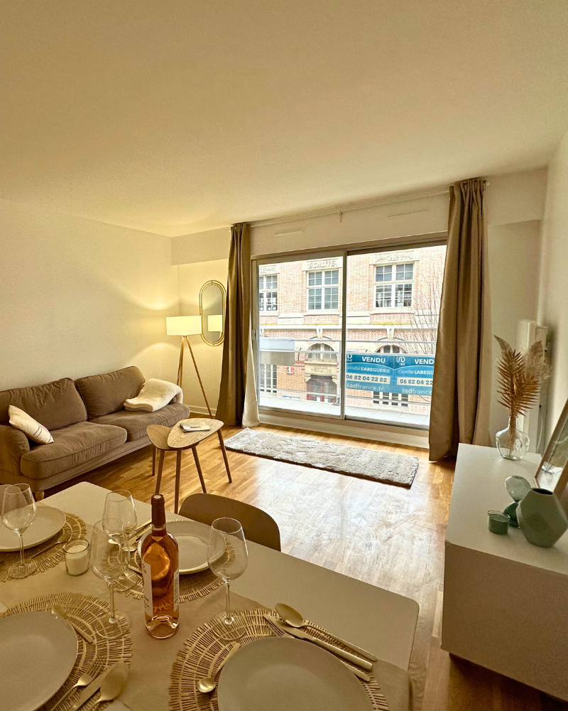 Un salon-salle à manger moderne avec un canapé marron, un lampadaire, de grandes fenêtres et une table dressée pour quatre personnes. On aperçoit les immeubles de la ville à travers la fenêtre.