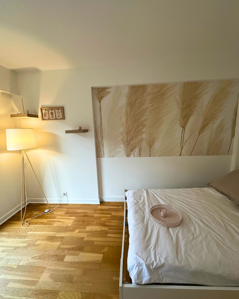 Une chambre minimaliste avec un lit, une literie blanche, un oreiller beige, un plateau avec une tasse, un lampadaire, une œuvre d'art murale représentant de l'herbe de la pampa et du parquet.