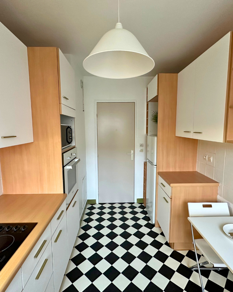 Cuisine étroite avec armoires en bois et blanches, sol à carreaux noirs et blancs, appareils électroménagers blancs et une suspension suspendue au plafond.