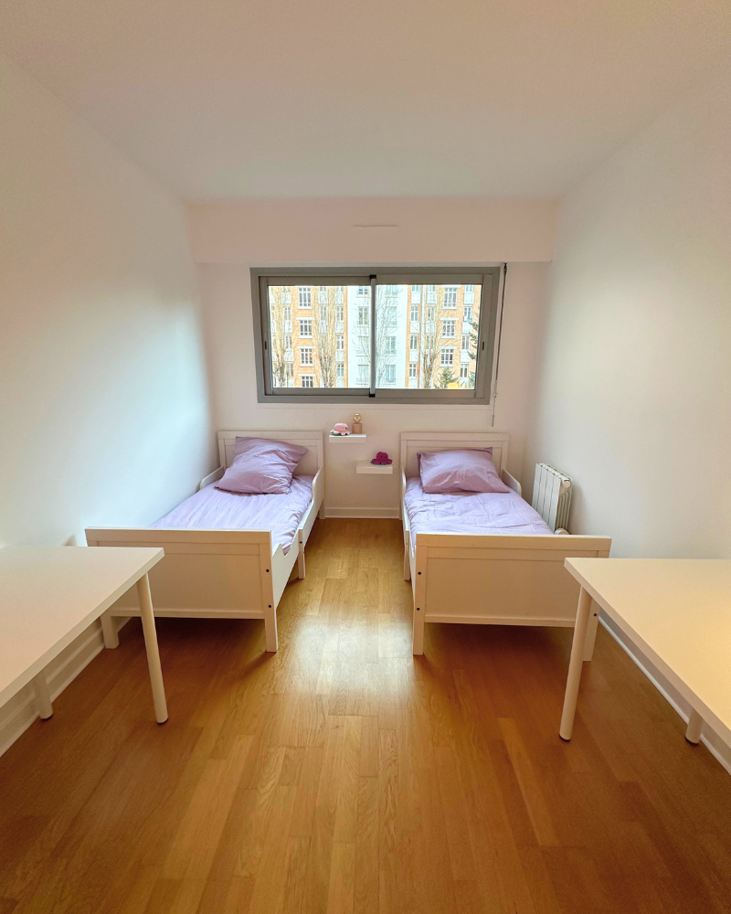 Deux lits simples avec une literie violet clair sont placés côte à côte dans une chambre lumineuse et minimaliste avec du parquet et une grande fenêtre donnant sur les bâtiments.