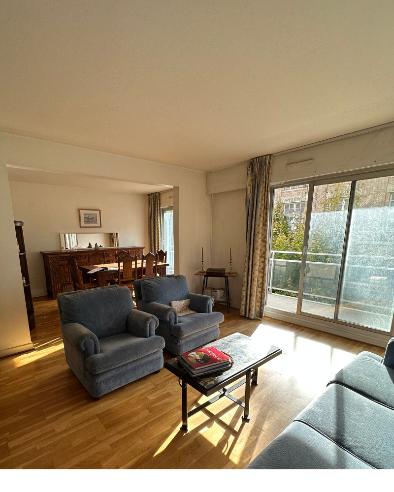 HS_2_avant_A Un salon avec des fauteuils bleus, un canapé, une table basse en verre et une table à manger en arrière-plan. De grandes fenêtres laissent entrer la lumière du soleil et offrent une vue sur un balcon et les bâtiments extérieurs.