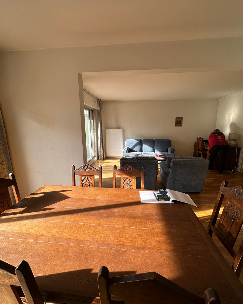 HS_2_avant_C Une salle à manger ensoleillée avec une table en bois au premier plan et un salon avec des canapés bleus et une personne travaillant à un bureau en arrière-plan.