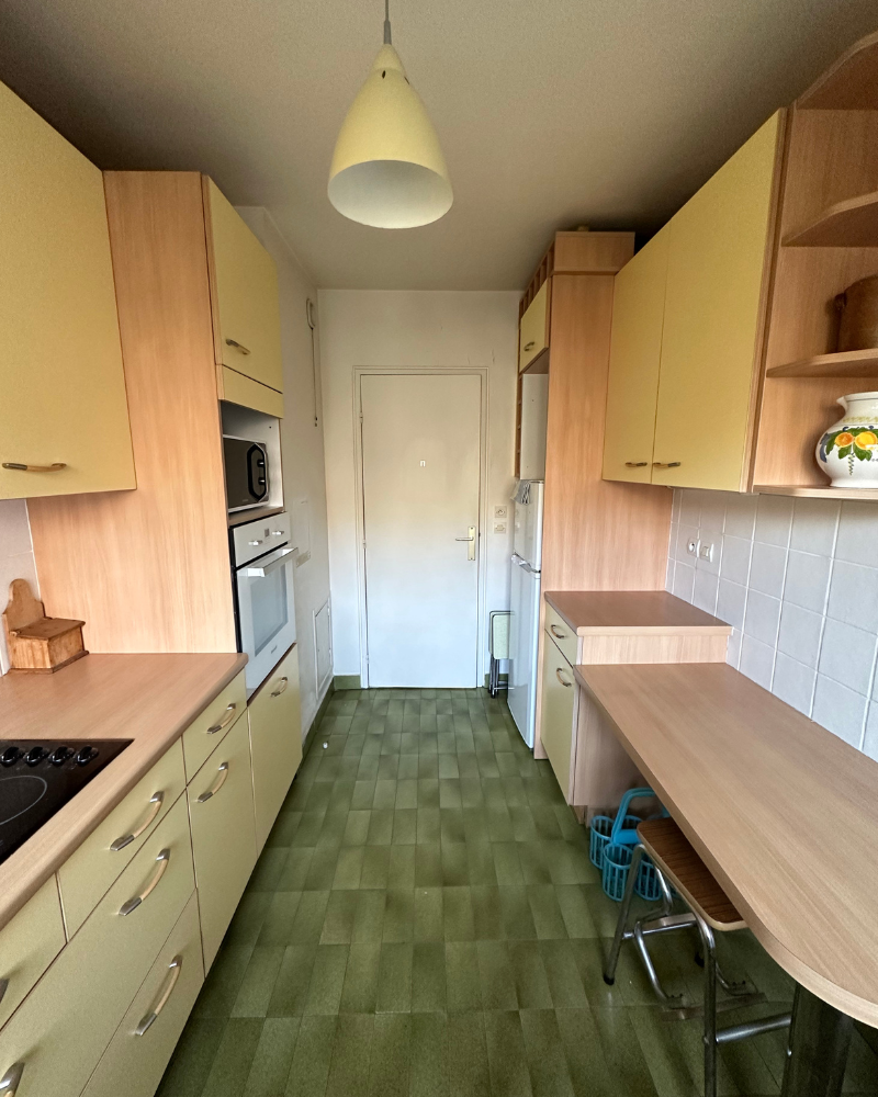 HS_2_avant_E Cuisine étroite avec placards en bois clair, sol à motifs verts, murs carrelés blancs, électroménagers intégrés et petit plan de travail avec deux tabourets. Une porte blanche se trouve au fond de la pièce.