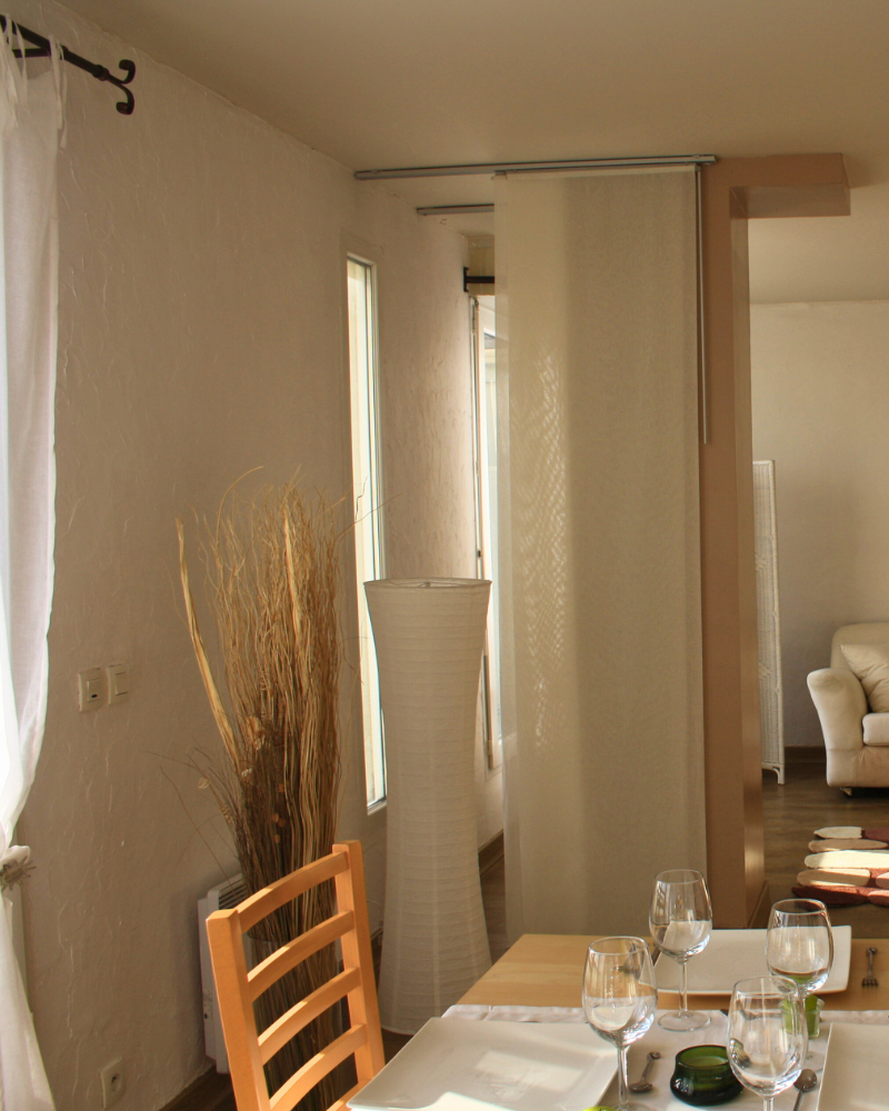 Au premier plan, un coin repas avec une chaise en bois et une table dressée ; à l'arrière-plan, on aperçoit un salon avec un canapé blanc, séparé par une cloison coulissante.