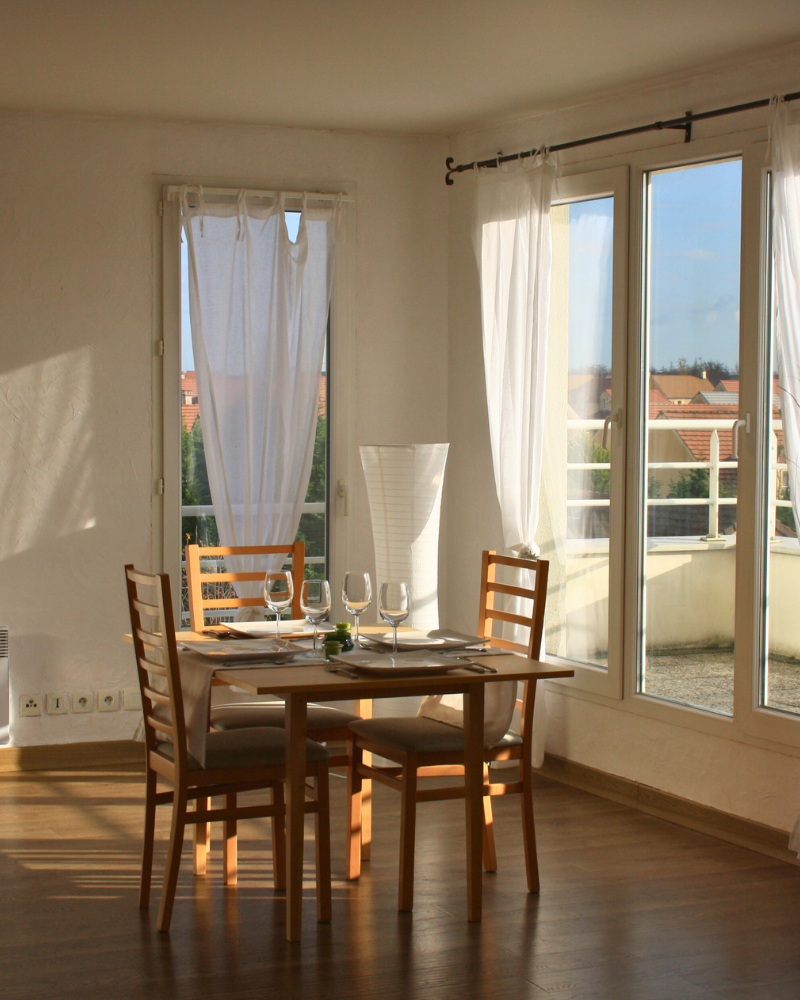 Une petite table à manger pour deux avec des chaises en bois, placée près de grandes fenêtres avec des rideaux blancs transparents, donnant sur un balcon et les toits extérieurs.
