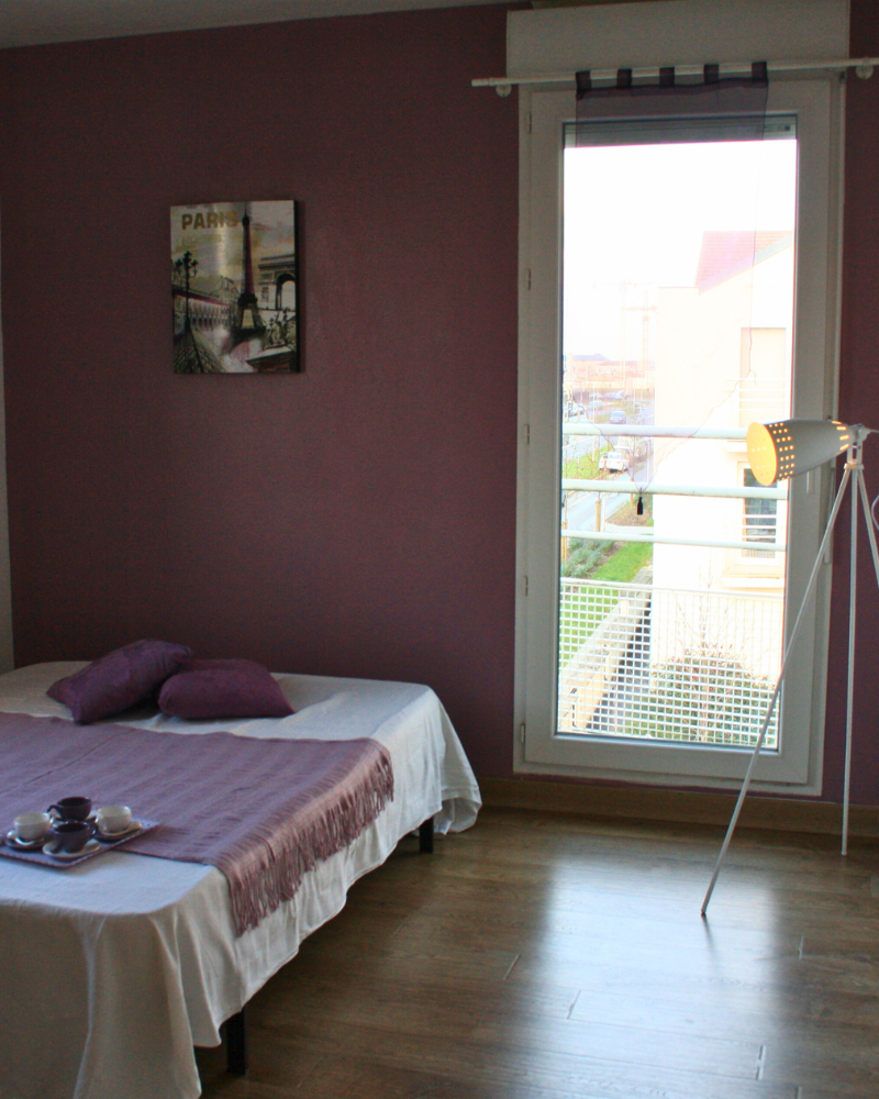 Chambre aux murs violets, un lit bas avec des oreillers et une couverture violets, une table d'appoint avec un service à thé, un lampadaire et une grande fenêtre montrant un bâtiment à l'extérieur.