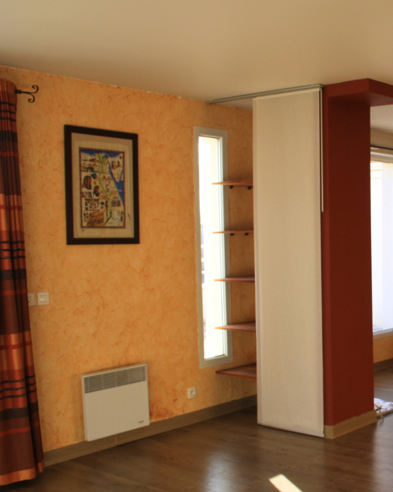 HS_3_Avant_A Une pièce avec des murs texturés orange, une œuvre d'art encadrée, une fenêtre verticale étroite, des étagères derrière un rideau blanc, un radiateur mural et des rideaux à rayures sur la gauche.