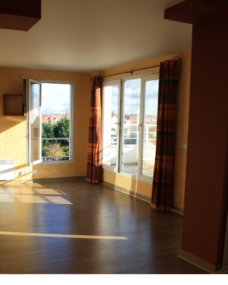 HS_3_Avant_C Une pièce lumineuse et vide, avec parquet, grandes fenêtres et rideaux à carreaux. La lumière du soleil entre à flots, projetant des ombres sur le sol. On aperçoit un balcon et des bâtiments à l'extérieur.