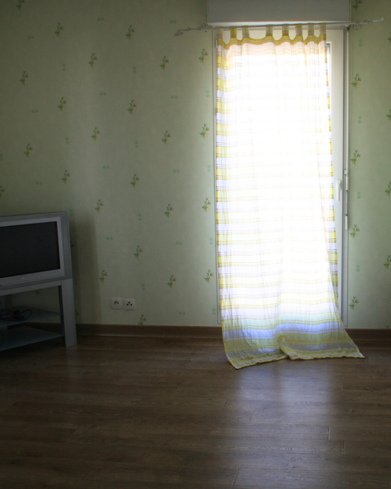 HS_3_Avant_E Une petite pièce presque vide avec du parquet, une télévision sur un meuble bas à gauche et une lumière filtrant à travers un rideau transparent à rayures recouvrant une porte vitrée.