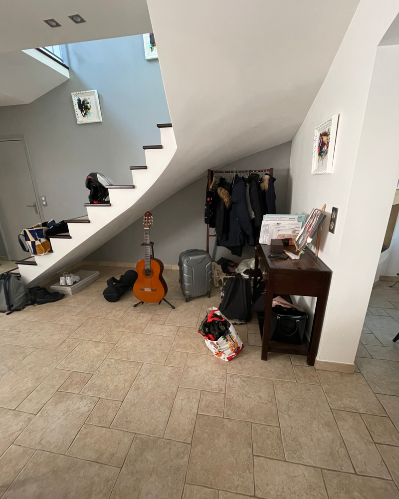 Rangement_sous_escaliers_1_avant Un couloir sous un escalier avec des manteaux sur un porte-manteau, une guitare, des valises, des sacs, des boîtes et une table avec des articles assortis, le tout sur un sol carrelé.