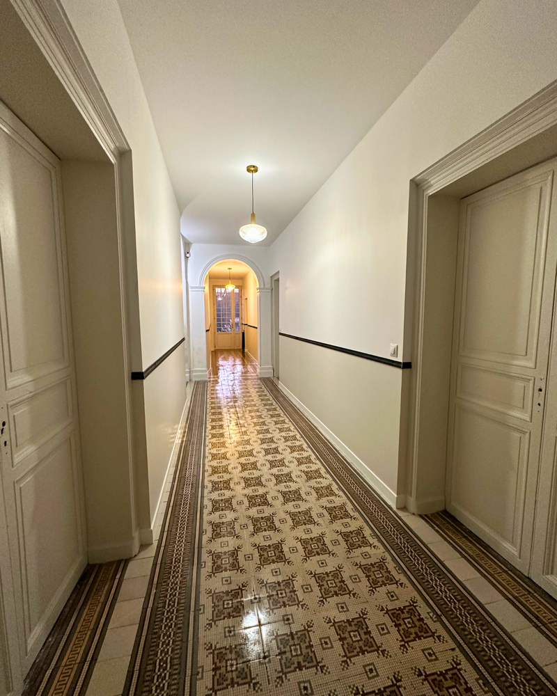 Un couloir faiblement éclairé avec du carrelage à motifs, des étagères et des meubles de chaque côté, menant à une porte avec des vitres au fond.