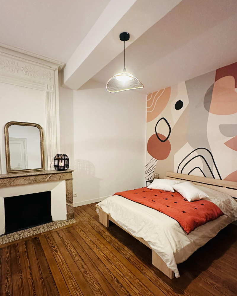Une chambre moderne avec un plancher en bois, un lit avec une literie blanche et rouge, un mur d'accent géométrique, une cheminée décorative, un miroir et une lampe sur le manteau.