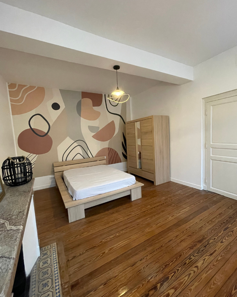 Une chambre minimaliste avec un cadre de lit en bois, un matelas, une armoire et une fresque abstraite sur le mur derrière le lit. La pièce est dotée de parquet et d'une suspension moderne.