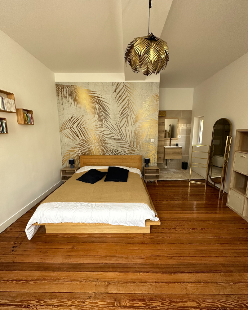 Une chambre minimaliste comprend un lit en bois avec une literie beige, un mur d'accent à motifs de feuilles d'or, du parquet et une décoration moderne, avec une salle de bain ouverte visible en arrière-plan.