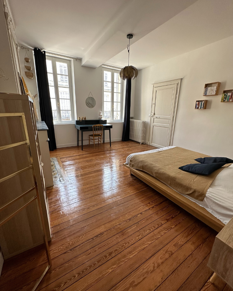 Pièce vide avec parquet, papier peint fleuri, deux grandes fenêtres, une porte fermée, un radiateur et un plafonnier suspendu.