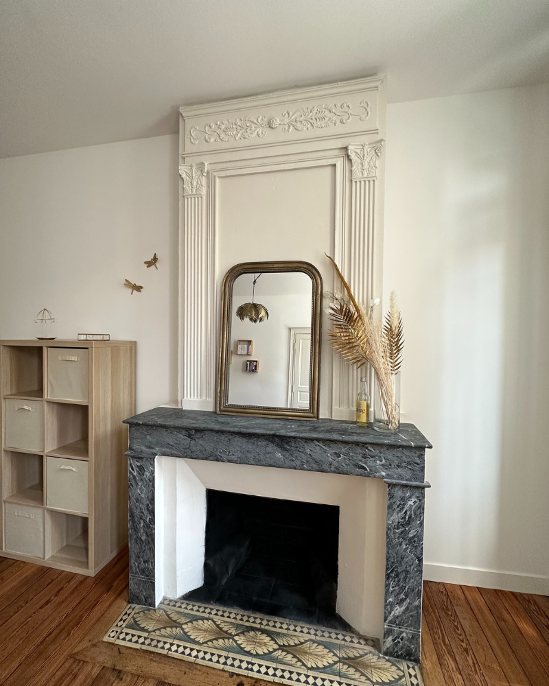 Pièce vide avec parquet, papier peint fleuri, porte blanche, cheminée en marbre et luminaire suspendu.