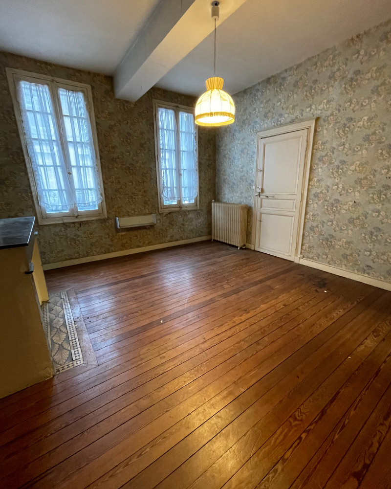 Une chambre avec parquet, un lit au premier plan, une cheminée à gauche et un bureau avec une chaise positionné entre deux grandes fenêtres avec des rideaux noirs.