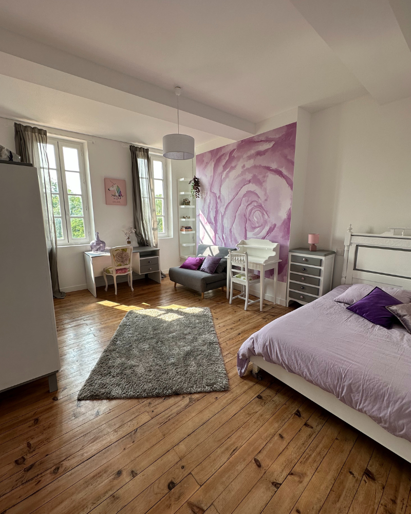 Chambre lumineuse avec parquet, lit, bureau, fauteuil et commode. Décoration lavande et blanc avec une grande fresque murale de roses sur un mur et lumière du soleil à travers de hautes fenêtres.