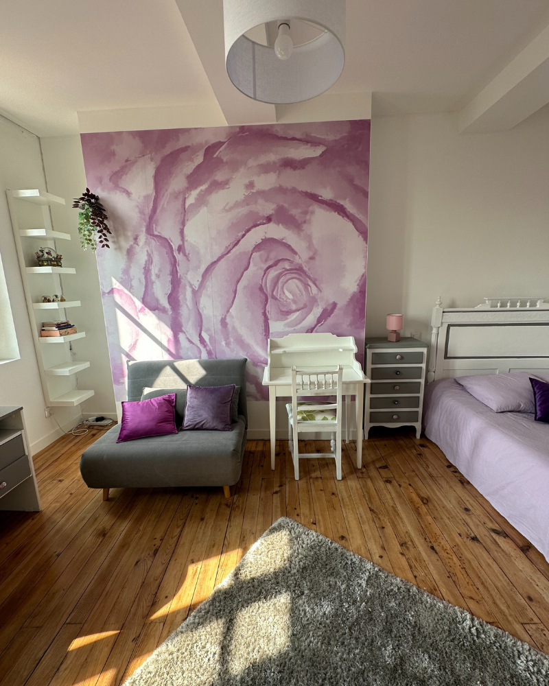 Une chambre lumineuse avec une fresque murale rose, une chaise longue grise, un petit bureau et une chaise blancs, un lit avec une literie lavande et du parquet.