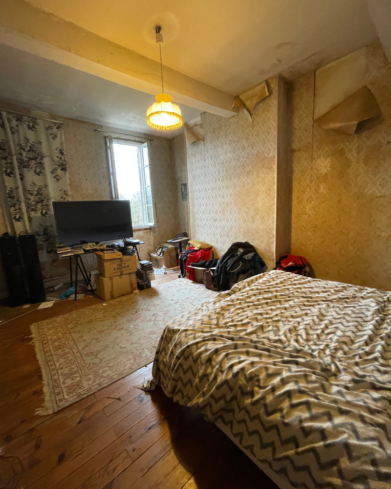 Une chambre avec un couvre-lit à motifs, une télévision sur une petite table, des boîtes, des sacs et du papier peint qui se décolle sous la lumière chaude d'un plafonnier.