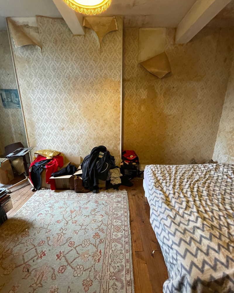 Une chambre encombrée avec du papier peint qui s'écaille, un tapis à motifs, un lit avec une couverture à chevrons et des vêtements et des cartons empilés contre le mur.