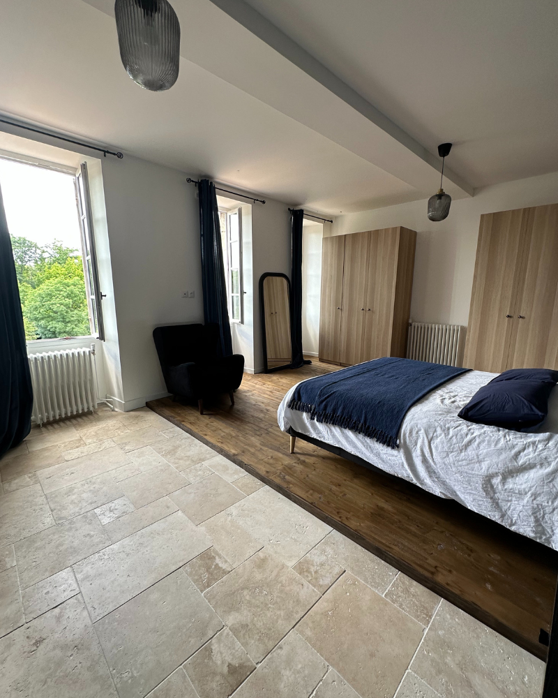 Chambre moderne avec un lit, un fauteuil, un miroir sur pied, deux armoires, du carrelage et du parquet, de grandes fenêtres et des rideaux sombres.