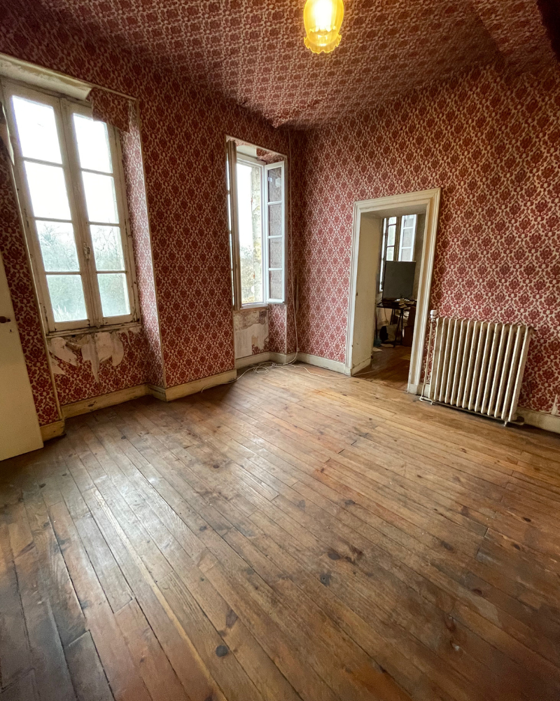 Une pièce vide avec du parquet usé, du papier peint à motifs rouges, deux grandes fenêtres, un radiateur et une porte ouverte menant à une autre pièce.