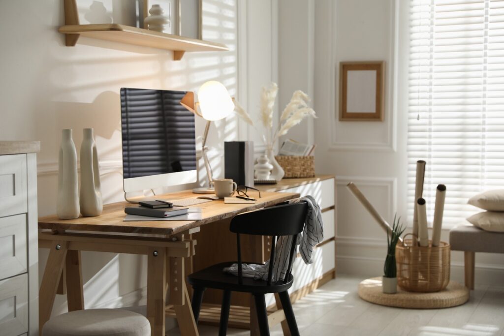 Un bureau à domicile moderne avec un bureau en bois, un ordinateur, une lampe, des livres, des objets de décoration et une chaise noire dans une pièce lumineuse avec de la lumière naturelle et des murs blancs.